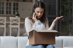 Dissatisfied angry woman, cheated client sit on sofa check received box, damaged or broken goods in parcel, talks to customers support, express complaints, looks annoyed. Bad delivery services concept