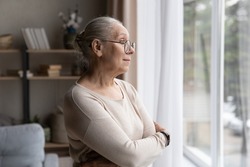 Older dreamy woman in glasses lost in thoughts looking pensive standing with arms crossed alone in living room staring out window, wait for grown up children, miss them spend time at home in solitude