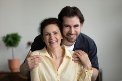 Grateful loving grown son embracing happy senior mom with tenderness, care, affection, looking at camera, smiling, laughing. Mature mother and adult child head shot portrait. Motherhood concept