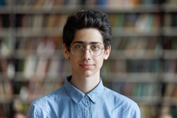 Head shot schoolboy guy posing in campus library on bookshelves background. 17s pupil in eyeglasses look at cam. Excellent student portrait, skill and knowledge, education, generation Z person concept