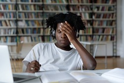 African student feels tired due to long preparations for college exam, deadline task, suffers from headache sit at table with laptop in library. Information overload during admission, stress concept
