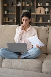 Serious thoughtful freelancer woman doing business paperwork at home, working laptop computer on couch. Renter girl paying domestic bills, taxes, using online app for online payment. Vertical shot