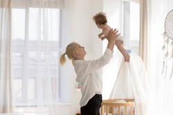 Happy cheerful new mom throwing adorable baby up in air, holding, lifting kid in arms, cuddling infant in diaper with love, care, enjoying motherhood, maternity leave, family leisure time at home