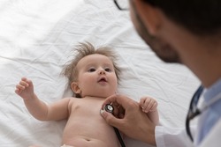 Kids doctor examining cute baby, applying stethoscope to chest, checking heartbeat, breath. Pediatrician visiting little infant patient lying on white sheet at home. Childcare, healthcare concept