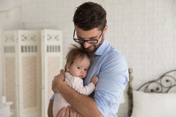 Peaceful happy handsome dad holding cute baby in arms, hugging little child with love, care, tenderness, hugging, ricking, calming daughter. Fatherhood, childcare, family concept
