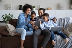 Cheerful young Black parents and curious little gen Z boy and girl resting together on home sofa, using laptop, tablet computer gadget, shopping internet, chatting online on social media