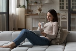 Happy cheerful reader girl enjoying novel story on couch, drinking hot beverage, reading paper book in cardboard cover, smiling, laughing, enjoying bookworms hobby, home leisure time