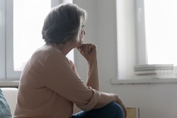 Serious older 50s woman looks out window spend time alone at home, feels sad or disappointed, waits or misses someone seated on sofa indoor. Loneliness, solitude on retirement, nostalgic mood concept