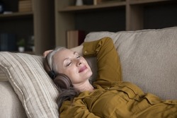 Peaceful carefree mature woman listening to music from wireless headphones, enjoying songs, lazy break, leisure time, chill, relaxing on comfortable sofa, meditating at home