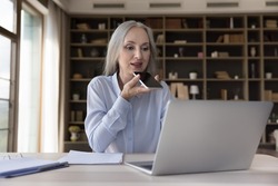Engaged mature business woman making phone call, talking on speaker at laptop on workplace table in home office, recording audio message, giving voice command to virtual assistant