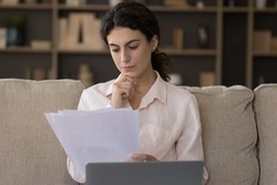 Serious focused young woman working with correspondence seated on sofa with wireless computer. Female holding papers, learn received formal document, analyze payment notification, do paperwork at home