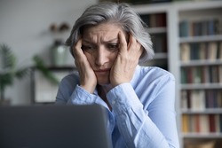 Frustrated senior retired woman touching head, looking at laptop screen with despair, getting bad news, reading online chat message, email letter. Problem with computer concept