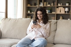 Attractive teenage girl sits on couch with smart phone. Young smiling woman spend time at home holding modern cellphone, share messages with friend, enjoy e-dating services, texting sms. Tech concept