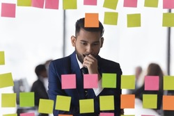 Thoughtful Indian businessman review notes written information on post-it stickers looks concentrated and concerned, deep in thoughts, search business ideas or solutions, makes tasks analysis concept