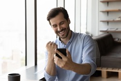 Happy excited businessman looking at smartphone screen, making winner yes gesture, smiling and laughing. Satisfied millennial man using mobile phone, reading good news, celebrating achieve, success
