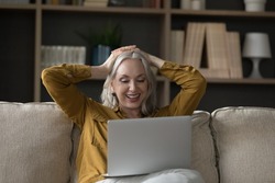Carefree relaxed older woman put hands behind head looks at laptop, enjoy new series of favourite movie resting seated on cozy sofa. Leisure and fun at home using modern tech, watch on-line TV concept