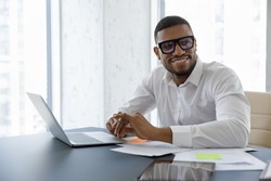 Handsome African businessman sit at desk with laptop smile staring aside in skyscraper office boardroom. Successful employee, career growth, modern tech, business owner portrait, aspirations concept