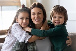 Headshot portrait of happy young Caucasian mother hug cuddle with cute small excited children at home. Loving caring two little kids embrace mom show affection and support. Family concept.