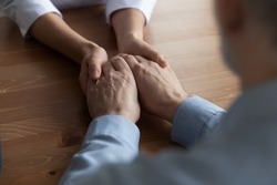 Close up young woman nurse holding hands of mature patient, sitting at wooden desk, doctor caregiver comforting lonely old aged man, expressing empathy and support, health problem and disease