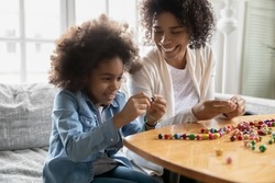 Smiling young African American mom or nanny sit at table play make bracelets with little girl child. Happy ethnic mother and small daughter have fun engaged in creative activity sting beads together