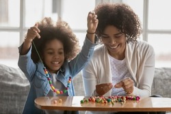 Happy young African American mom have fun string colorful beads play with little ethnic daughter. Excited mother and small girl child involved in creative activity, make bracelets together
