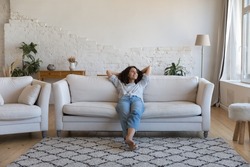 Happy thoughtful young Latin woman resting on comfortable couch at home, looking away, smiling, dreaming, thinking, relaxing, enjoying leisure time, breathing fresh air