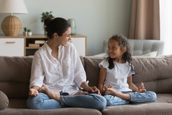 Cheerful Indian mom teaching girl to do yoga at home, sitting in lotus pose on couch, talking to daughter kid, laughing, smiling, discussing meditation, enjoying activity, exercises, family leisure