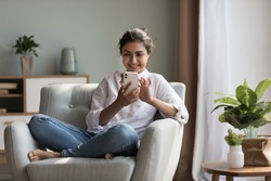 Happy pretty millennial Indian girl relaxing at home, resting in armchair, typing on smartphone, using online app, software, shopping on Internet, making video call. Mobile phone communication