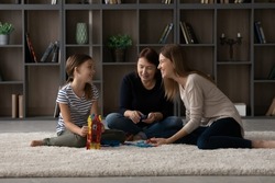 Happy three female generations family sitting on floor carpet, having fun playing toys entertaining together in modern living room, joyful preteen girl entertaining with mommy and granny on weekend.