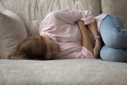 Depressed unhappy ill young woman lying on couch in embryo pose, suffering from belly ache, spasm, miscarriage symptoms, feeling strong pain. Health problems, emotional crisis