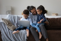 Happy caring young African American father using laptop with adorable little children son daughter, sitting on couch at home, having fun playing games online and communicating at home, tech addiction.