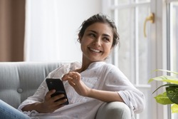 Peaceful Indian woman relaxing on cozy chair with smartphone, smile staring aside enjoy carefree weekend and modern wireless tech. Making call, ordering goods and services on-line, e-date apps usage