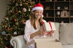 Joyful sincere young woman in festive hat taking wrapped gift from big carton box, feeling excited of getting Christmas present, satisfied with quick delivery service, New Year celebration concept.