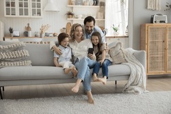 Cheerful mom, dad and two little kids using mobile phone at home together, hugging on sofa, playing online virtual game, taking self video for blog, social media, making family call