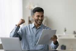 Positive Indian professional man celebrating business success, achievement, victory, holding paper document, letter, notice, contract, agreement, offer, making winner yes hand