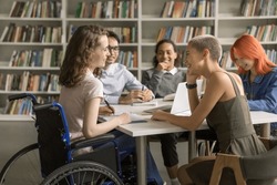 Young student girl with disability cooperating on class project in library with college friends, using wheelchair, sitting at table, talking, chatting with classmates, laughing
