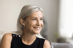 Happy mature blonde lady looking away with toothy smile, thinking, dreaming, posing at home, laughing in good thoughts. Positive senior woman close up candid facial portrait