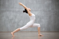 Side view portrait of beautiful young woman wearing white tank top working out against grey wall, doing yoga or pilates exercise. Standing in Warrior one pose, Virabhadrasana. Full length