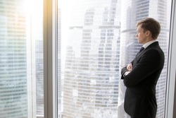 Successful businessman in classical formal suit standing in office with hands crossed on chest, looking through window at big city skyscrapers, thinking of new deals, dreaming. Copy space for text