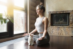 Young attractive smiling woman practicing yoga, sitting in Half Lotus exercise, Ardha Padmasana pose, working out, wearing sportswear, meditation session, indoor full length, home interior, cat near