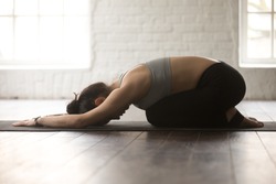 Young attractive sporty yogi woman practicing yoga concept, sitting in Child exercise, Balasana pose, working out, wearing black sportswear, full length, white loft studio background, side view