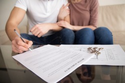 Close up of tenants signing rental agreement, renters couple sitting on couch, male hand with a pen putting signature, focus on document and keys, joint residential tenancy, lease contract 