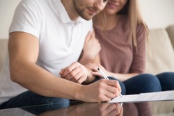 Close up of couple signing documents, young man putting signature on document, his wife sitting next to husband holding his arm, real estate purchase, first time home buyers, prenuptial agreement 