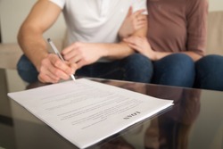 Close up of signing loan agreement, couple sitting on sofa, male hand with a pen setting signature, taking bank credit with easy terms of payment and low interest rate for purchasing property 