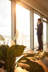 Successful businessman starting working day with cup of coffee, standing in modern sunny office interior, looking through full-length window at big city, having break, deep in thoughts, vertical view 