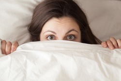 Funny surprised girl covering half of face with white blanket, young scared woman hiding and peeking from duvet, afraid of night monsters, feels embarrassed, wide awake, head shot close up, top view