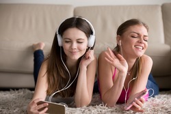 Two happy girlfriends in stylish white earphones and headphones lying on soft carpet on floor, sisters enjoying their favorite music tunes on modern smartphones. Siblings having fun at home concept. 