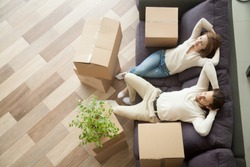 Couple resting on couch after moving in, man and woman relaxing on sofa just moved into apartment with cardboard boxes on floor, happy satisfied homeowners enjoying first day in new home, top view