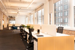 Workstations sprawl across a modern,  open concept office building. Natural light fills the room from large windows along the walls.