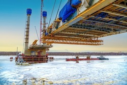 Construction of a new road bridge continues across the Sheksna river. Cable-stayed bridge in Cherepovets at sunset. Fragment of the unfinished construction of the city highway.Cargo barge on ice. 